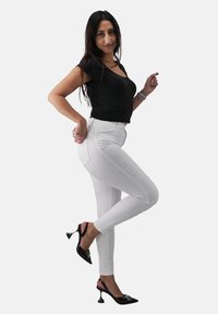 Jeans skinny bianchi abbinati a una maglia nera aderente e scarpe con tacco nere con dettagli argento. La modella posa con una gamba sollevata.