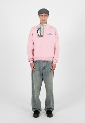 Man draagt een roze trui met het logo "Prohibited Racing Club", een geruite nek sjaal, een grijze platte pet, losse verweerde spijkerbroek en zwarte schoenen, staand in een rechte houding.