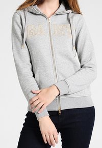 Sudadera gris con cremallera, con "GANT" bordado en dorado, dos bolsillos delanteros, capucha con cordón y puños acanalados. Hecha de tejido suave y cómodo.
