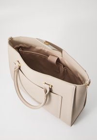 Beige rakenne käsilaukku, joka on valmistettu teksturoidusta materiaalista, jossa on kultainen varustus, ylhäällä oleva kahva ja tilava, vuorattu sisätila, jossa on tasku.