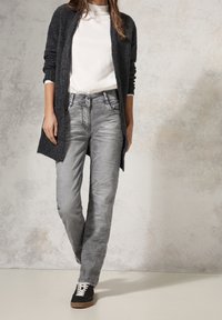 Cardigan grigio in lana sopra una camicia bianca, abbinato a jeans grigio chiaro con una leggera lucentezza e due bottoni in vita, completato da sneakers nere.
