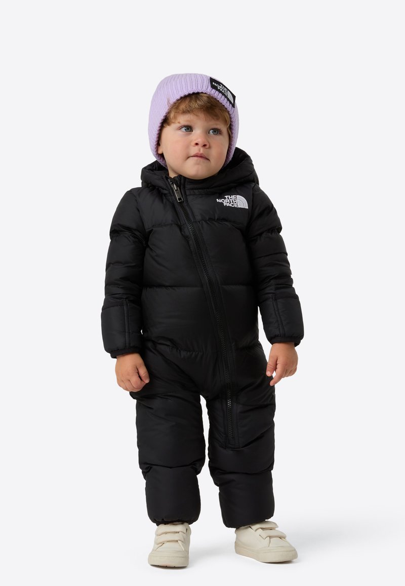 Black padded snowsuit with zip front and logo detail, paired with a lavender beanie. Cream-colored shoes with strap accents.