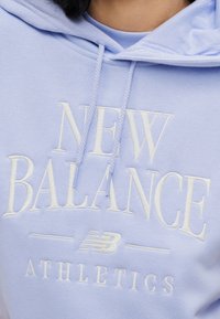 Ανοιχτό μπλε φούτερ με κουκούλα με λευκή κεντημένη επιγραφή που γράφει "NEW BALANCE ATHLETICS." Διαθέτει κουκούλα με κορδόνι και απαλό υφή.