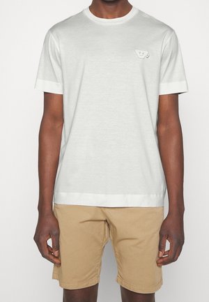 Mężczyzna ubrany w prostą białą koszulkę z krótkim rękawem z małą naszywką na piersi oraz beżowe spodenki do kolan.