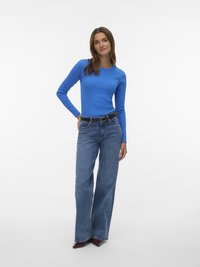 Woman standing, wearing a bright blue long-sleeve top, high-waisted wide-leg jeans with a black belt, and pointed dark shoes.