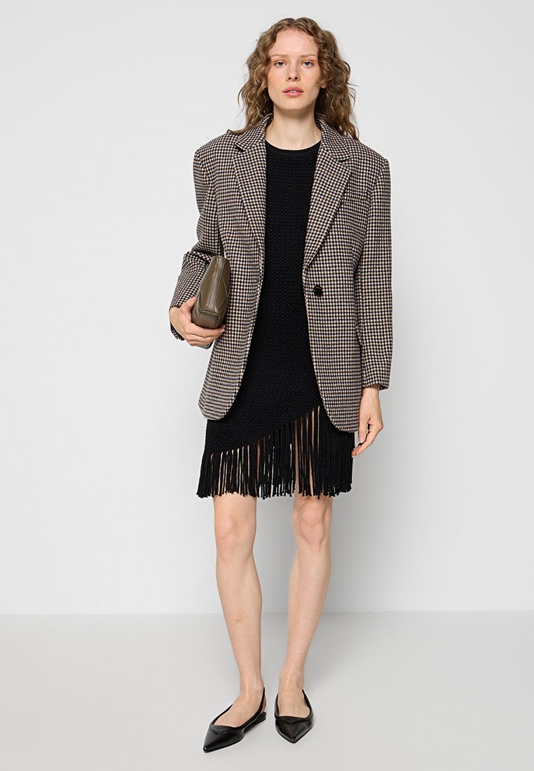 Braun-schwarz karierter Blazer über einem schwarzen Strickkleid mit Fransen am Saum. Das Model hält eine braune Clutch und trägt schwarze spitze Ballerinas.