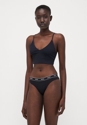 Black bralette with thin straps and V-neck, paired with black bikini bottoms featuring a white "HUGO" logo on the waistband.