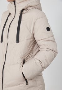 Beige polstret jakke med hette, sorte glidelåser og stropper. Har sidelommer og en rund svart logo på venstre erme.