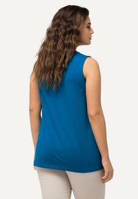 Top senza maniche, con scollo rotondo, realizzato in tessuto teal solido, caratterizzato da una superficie liscia e una lunghezza lunga. Modello visto di spalle che indossa pantaloni beige.