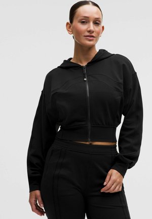 Kobieta ubrana w czarną krótka bluzę na zamek z długimi rękawami oraz dopasowane czarne spodnie, stojąca na jednolitym jasnym tle.