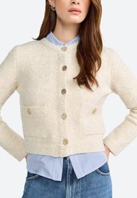 Cardigan crema realizzato in tessuto testurizzato, con bottoni color oro e due tasche anteriori, indossato sopra una camicia a righe blu e bianca.