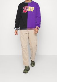 Czarna i purpurowa bluza dresowa z haftowanym logo, ściągaczami na rękawach i paskowanym obszyciem na dole. Beżowe sztruksowe spodnie i ciemnozielone trampki.