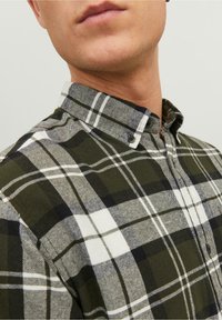 Camicia a quadri verde e bianca con bottoni e collo con revers, caratterizzata da una texture morbida e bottoni scuri su uno sfondo oliva.