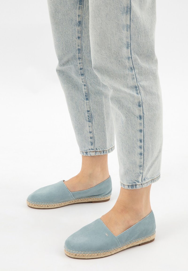 Espadrillas azzurro chiaro slip-on con suole avvolte in juta indossate con jeans a caviglia a lavaggio chiaro su sfondo bianco.