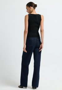 Top de tirantes negro ajustado con textura brillante, combinado con unos jeans de tiro alto de campana azul oscuro. La modelo es vista desde la parte posterior.