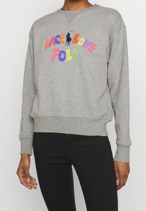 Personne portant un sweat-shirt gris avec un texte coloré "Peace Love Polo" et un logo joueur de polo noir, associé à un pantalon noir.