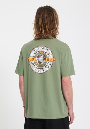 Persoon met schouderlang haar die een groen T-shirt draagt met een Volcom Stone Co-graphic met een wereldbol en de tekst "1991 Earth Tripper".