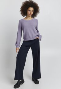 Lila gestrickter Pullover mit weitem Rundhalsausschnitt, kombiniert mit navyblauen weiten Hosen. Strukturiertes Material und lässige Passform. Schwarze klobige Schuhe.
