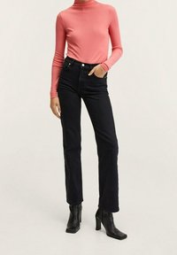 Femme portant un haut long en corail ajusté, un jean noir taille haute et des bottines à talons noires, se tenant avec une main dans la poche.