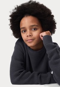 Maglione grigio scuro a maglia con un motivo a nido d'ape testurizzato e colletto a costine. Un bambino si inclina in avanti, appoggiando il mento sulla mano, con i capelli ricci.