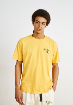 Tricou din bumbac galben, cu mâneci scurte, având un decolteu rotund și un mic logo negru imprimat pe partea stângă a pieptului.
