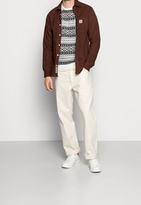 Ruskea napillinen paita valkoisen kuvioidun villapaidan päällä, yhdistetty vaaleisiin beigeihin housuihin ja valkoisiin lenkkareihin. Kangasten pinnat ovat huomionarvoisia.