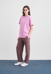 Roosa puuvillane t-särk valge logoga, pruuni logoga dressipükstega ja valgete tossudega sinise vaibaga põrandal.