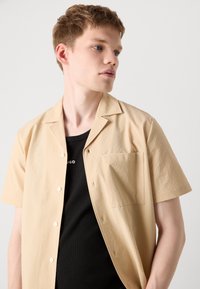 Kurzärmliges, hellbeiges Hemd aus strukturiertem Stoff, mit Knopfleiste und aufgesetzter Brusttasche. Tragen über einem schwarzen Top mit rundem Ausschnitt.
