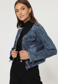 Chaqueta de denim con un acabado azul desgastado, cierre de cremallera en la parte delantera, dos bolsillos en el pecho y detalles en cremallera de tono plateado. Combinada con un suéter negro de cuello alto.