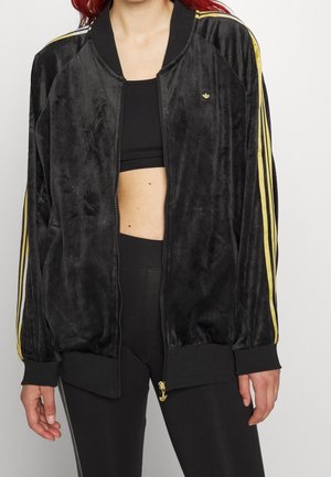 Osoba nosząca czarną welurową kurtkę Adidas ze złotymi paskami i logo, na czarnym staniku sportowym i czarnych legginsach.