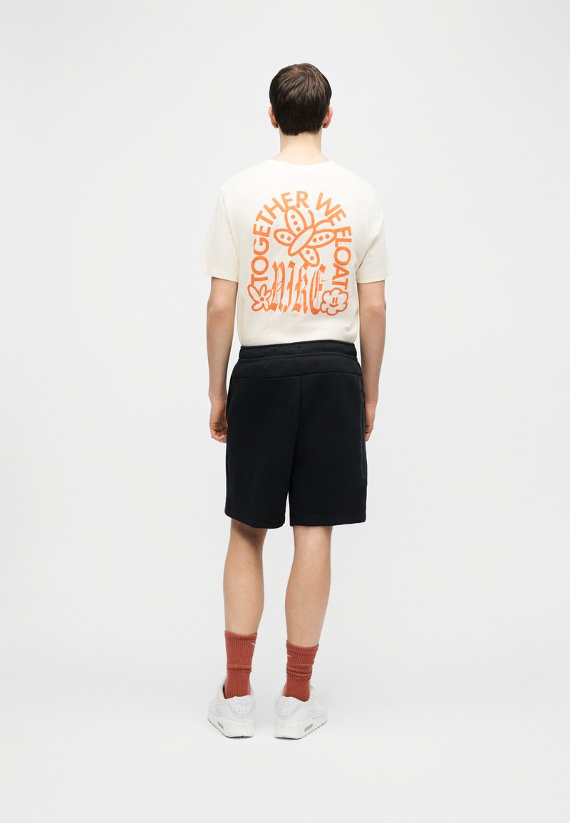 Homme debout, de dos, portant un T-shirt blanc avec un papillon orange et le texte « Together We Float », un short noir, des chaussettes couleur rouille et des baskets blanches.