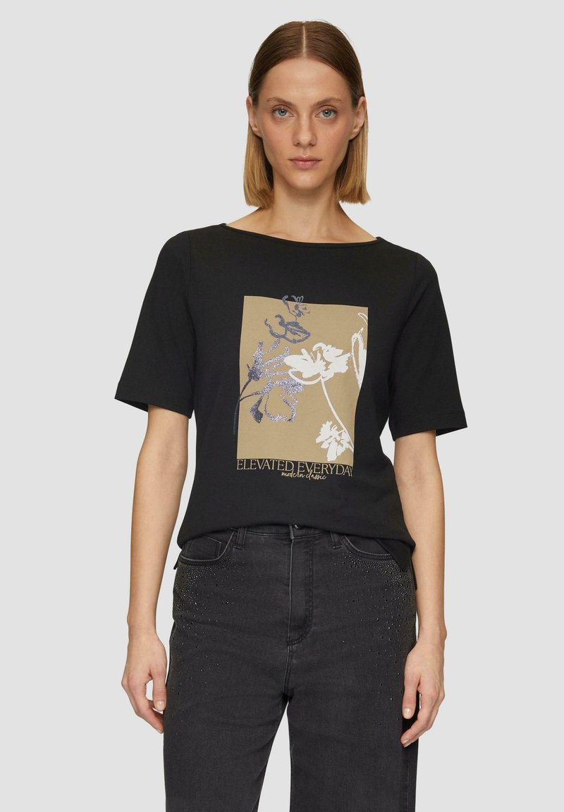 T-shirt noir à manches courtes avec un motif floral central crème et beige et un design de texte "ELEVATED EVERYDAY". Texture de tissu lisse.
