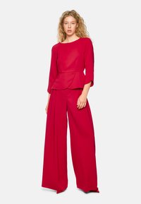 Rotes Peplum-Oberteil mit Dreiviertelärmeln, kombiniert mit weit geschnittenen Hosen, beide aus einem glatten Stoff mit maßgeschneiderter Passform.