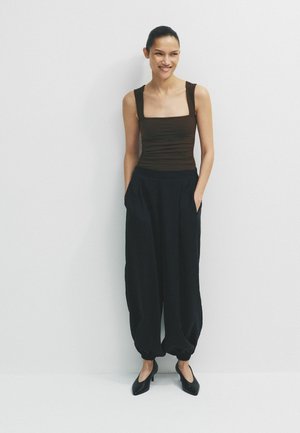 Woman standing against white wall wearing sleeveless brown top, loose black pants with gathered cuffs, and black heels, hands in pockets, smiling.