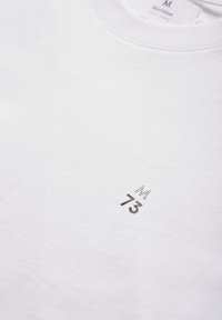 Balta medvilninė marškinėlių su apvalia iškirpte, ant kurių apatinėje dešinėje pusėje yra mažas juodas spaudinio dizainas su skaičiumi "73".