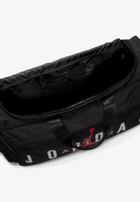 Sort duffel bag med en lynlåsåbning, der har et stort trykt logo, flade håndtag og en lille inderlomme. Tekstureret stof.