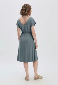 Türkiser Kurzarmkleid aus weichem Stoff, mit tiefem V-Ausschnitt im Rücken und elastischem Bund, das bis zum Knie fällt. Getragen mit beigen Sandalen.