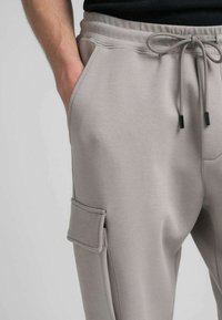 Pantaloni della tuta grigio chiaro realizzati con materiale morbido, dotati di vita elasticizzata con coulisse, tasche laterali e una tasca in stile cargo sulla coscia.
