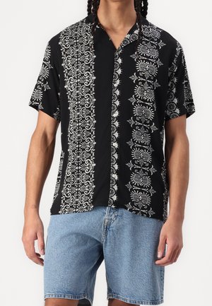 Homme portant une chemise noire à manches courtes avec des motifs floraux blancs et en spirale, associée à un short en jean bleu clair.