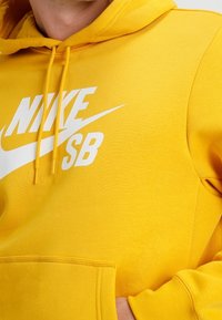 Gul Nike SB hoodie med vit logotyp, snoddar som hänger, och en framficka med en hand i.