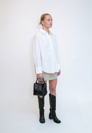 Donna con capelli biondi con una camicia bianca oversize, gonna beige, stivali neri al ginocchio, che tiene una borsa nera, in piedi contro un muro bianco.