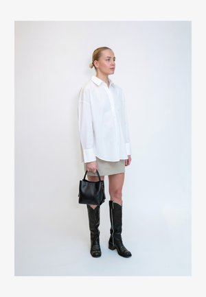 Femme aux cheveux blonds dans une chemise blanche oversize, jupe beige, bottes noires montantes, tenant un sac à main noir, debout contre un mur blanc.