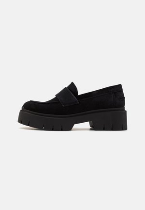 Musta mokkanahkainen loafer paksulla kumipohjalla ja matalalla korolla, slip-on-tyyli leveällä nauhalla päältä.