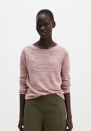 Person mit kurzen Haaren, die einen hellrosa Langarmpullover und olivgrüne Hose vor einem schlichten weißen Hintergrund trägt.