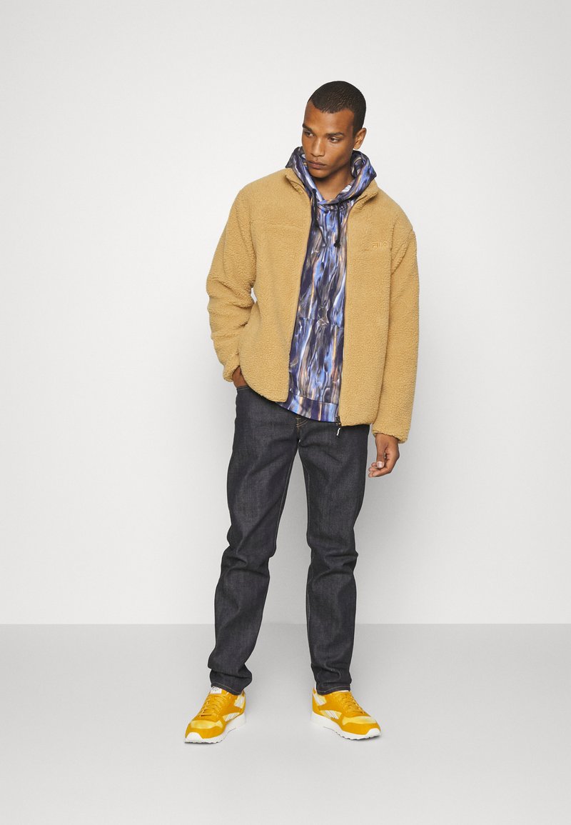 Giacca di pile beige con zip sopra un felpa blu a fantasia, abbinata a jeans scuri e sneaker gialle brillanti. Texture liscia e vestibilità rilassata.