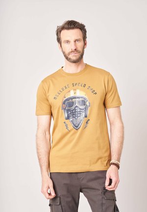Man met baard die een mosterdkleurig T-shirt draagt met een vintage helm en bril erop en donkere cargobroek tegen een effen achtergrond.