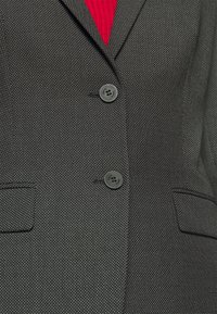 Sort blazer med en tekstureret stof og et subtilt mønster, to knapper og to forlommer. Rød tekstil synlig nedenunder.