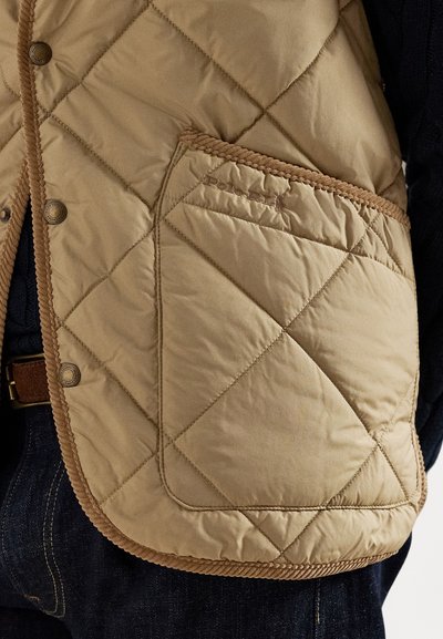 Beżowa pikowana kamizelka w diamentowy wzór, z brązowymi akcentami i boczną kieszenią. Napowe zapięcia i kontrastowe obszycia wzdłuż krawędzi.