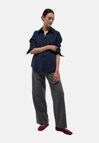 Chemise bleu marine à boutonner avec des manches retroussées, associée à un pantalon large gris et des chaussures rouges texturées. Design minimaliste, tissu lisse.