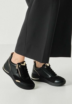 Person trägt schwarze Sneakers mit goldenen Akzenten und Reißverschlüssen, kombiniert mit schwarzen Hosen mit weitem Bein auf hellem Boden.